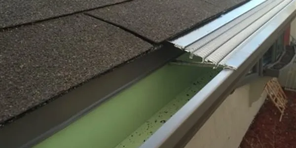 Close-up view of a roof with asphalt shingles and metal gutter guard, partially detached and bent, exposing the green interior of the rain gutter—an issue that may require General Contracting expertise to prevent damage to siding or windows.