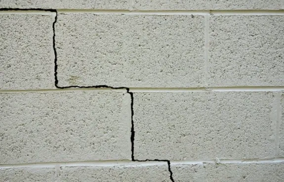 A close-up of a white concrete block wall with large, jagged cracks running horizontally and vertically, suggesting potential issues with the building’s siding or gutters.