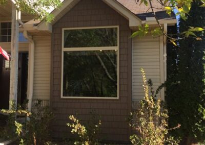 A house with beige siding and brown roofing features a large front window. Bushes line the window, bordered by bricks, with scattered leaves on the ground. Tree branches frame the scene under a clear blue sky.