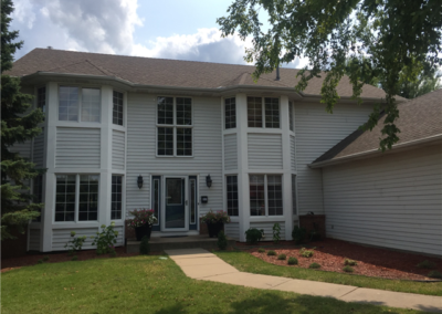 Two-story suburban house with light gray siding, large windows, and a teal front door. The yard features green grass, small plants, and a curved sidewalk. Quality windows and expert general contracting add to its curb appeal. Trees and a cloudy sky in back.