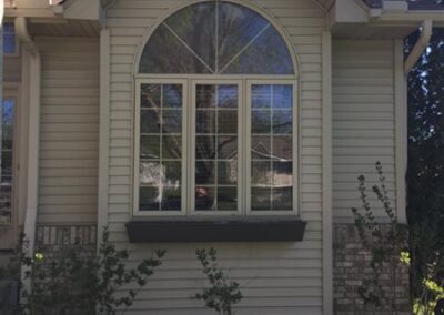 A large arched window with white trim is set in the beige siding of a house. Below, a bed of small rocks features bushes and garden statues. Clean gutters line the roof, all beneath a clear blue sky.