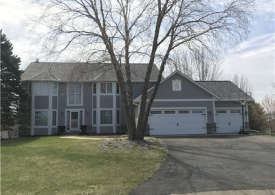 A two-story gray house with white trim in Woodbury features large windows, a three-car garage, and well-maintained gutters. A bare tree stands in front, a basketball hoop is by the driveway, and the partly green lawn sits under cloudy skies.