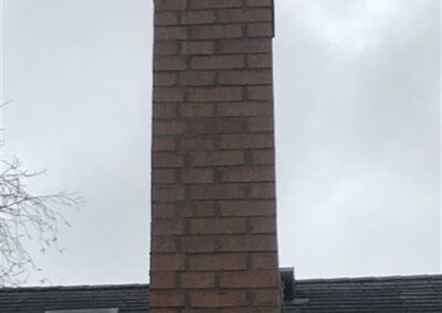 A red brick chimney with a metal flue extends above a shingled roof and nearby gutters, set against a cloudy sky. The chimney leans noticeably to one side.
