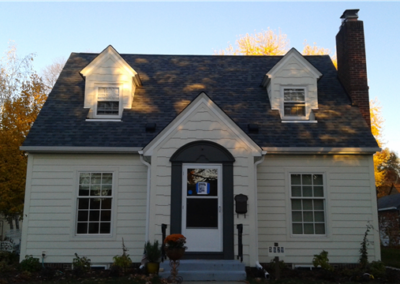 A small cream-colored house with dark shingles, two dormer windows, and a central chimney showcases quality roofing. The tidy front yard is framed by autumn trees, making this home a charming example of expert general contracting.
