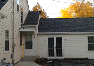 A light-colored house in Woodbury features dark gray shingles, white trim, and two sets of French doors. Trees with autumn leaves stand in the background, and the ground near the windows is covered with black plastic.