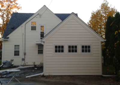 A cream-colored house with new siding and a smaller attached structure, possibly a garage, featuring three rectangular windows. Patio furniture and some construction materials are visible in the foreground. Trees with autumn foliage surround the house.