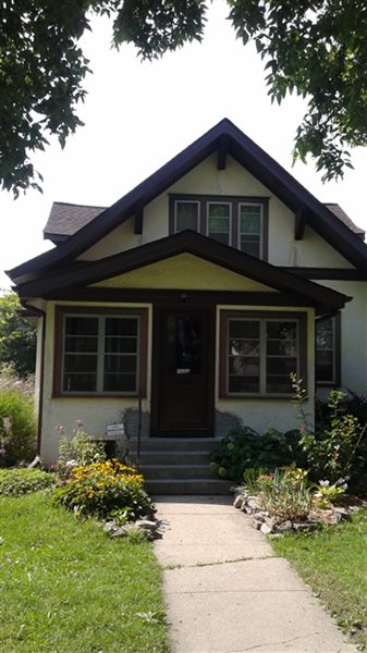 A small, two-story Woodbury house with a front porch, brown trim, and cream-colored exterior. Stone steps lead to the porch, bordered by a flower garden. New windows frame lovely views of trees and lawn in the foreground.
