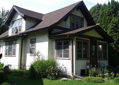 A two-story house in Woodbury with white exterior walls, brown trim, and a dark shingled roofing, surrounded by green grass, shrubs, and flowering plants on a sunny day.