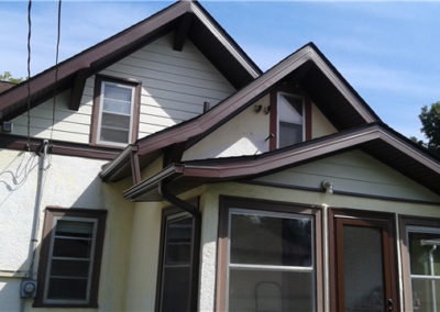 A close-up view of the exterior siding and roofing of a beige house with brown trim in Woodbury, featuring multiple gabled roofs and several windows under a clear blue sky. Some trees are visible in the background.