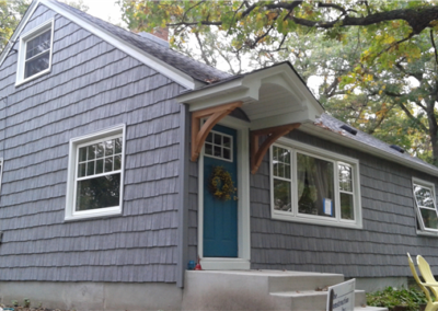 A gray shingled house with white trim, new siding, a blue front door with a wreath, and a small porch. Two yellow chairs are on the right side, and trees with green leaves surround the home’s windows.