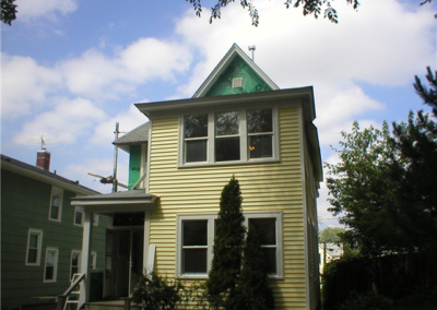 A two-story yellow house with white trim and a pointed roof, partially under renovation with exposed green siding. A tall bush stands in front, and the sky is partly cloudy.