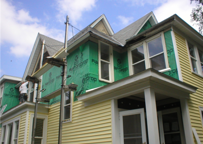 A two-story house is being renovated, with green weatherproof wrap on the upper exterior walls and new yellow siding on the lower walls. Scaffolding is set up for workers installing both siding and gutters. The sky is partly cloudy.