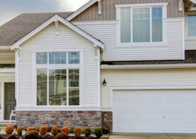 Two-story suburban house in Woodbury with white siding, stone accents, large windows, and a double garage. The front yard features neatly trimmed bushes, flowers, a concrete driveway, and well-maintained roofing and gutters.