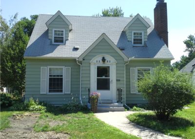 A small, light green house with a steep gray roofing, two dormer windows, a central white door, a tall brick chimney, and some bushes and plants in the front yard on a sunny day.