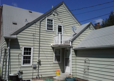 A light green Woodbury house with gray roof shingles and siding features a small white balcony above a back door, visible utility wires, garden tools, and a yellow bucket on a concrete patio, all under a clear blue sky.