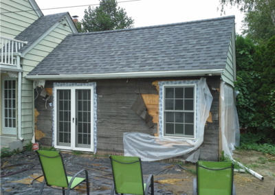 A small house or shed is under renovation, with exposed insulation and partially installed siding. Two windows and a door are framed, plastic sheets cover the ground, and three green chairs sit in the foreground—general contracting is underway.