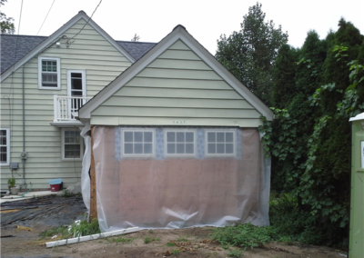 A detached garage with cream siding is under renovation by Woodbury General Contracting. The garage door opening is covered with clear plastic and plywood, while windows await installation. Bare dirt and construction materials are visible around.