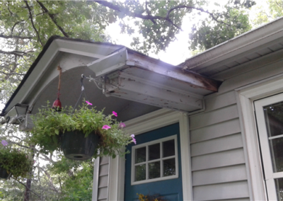A close-up of a small porch roof over a blue door with hanging flower baskets in Woodbury. The roof shows visible water damage and rot along the edge. Green trees, part of a window, and siding are visible.