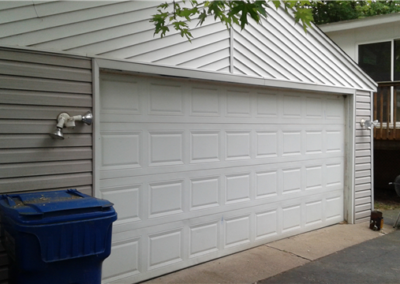 A closed white garage door on a house with gray siding, two blue recycling bins in front, and a black charcoal grill near a wooden deck railing—showcasing quality craftsmanship and skilled siding work.