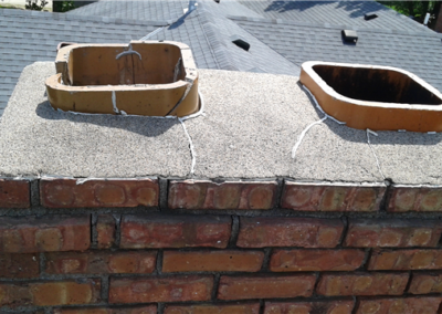 A close-up view of a brick chimney with two rectangular clay flue liners on top, each secured with white string, on a shingled roof. The chimney surface shows signs of wear, with siding and windows visible in the background.