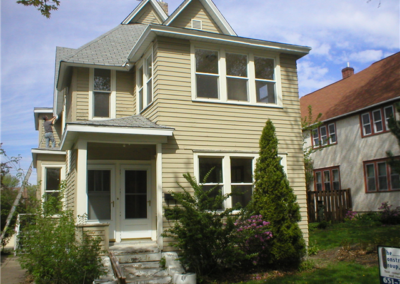 A two-story beige house with white siding and trim, featuring large windows and a small covered porch, is surrounded by shrubs and trees beside another house on a residential street under a blue sky.