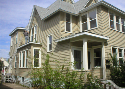 A person stands on a ladder painting the upper trim and siding of a large, beige house with multiple windows and a front porch. Some plants grow along the foundation near the gutters. It appears to be a sunny day.