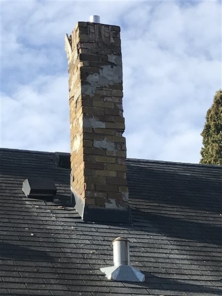 A brick chimney with visible damage and uneven bricks stands atop a shingled roof under a partly cloudy sky in Woodbury. A small metal vent and well-maintained gutters are also visible on the roof in the foreground.