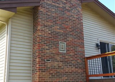A tall rectangular red brick chimney attached to the exterior of a light-colored house with vertical siding and modern windows, next to a second-story wooden deck with railing.
