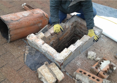 A person wearing gloves works on roofing repairs, rebuilding a brick chimney on a roof in Woodbury. Bricks and chimney components are scattered nearby, and the old chimney flue lies on its side.