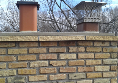 A brick chimney with two metal chimney caps, one brown and one silver, stands tall against leafless trees and a cloudy sky—showcasing quality roofing commonly seen in Woodbury.