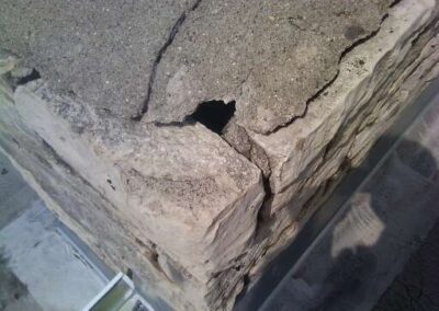A close-up view of a damaged concrete and stone structure corner in Woodbury, showing a deep crack and a hole in the top surface near the gutters, with visible wear and exposed edges.