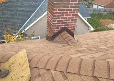 A brick chimney rises from a shingled roof, with metal flashing and shingles installed around its base. Nearby rooftops, siding, windows, trees, and a yard complete the background scene.