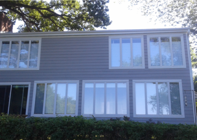 A two-story gray house with large rectangular windows on both floors, surrounded by greenery and shaded by tall trees. Expert siding work enhances the exterior as sunlight filters through the leaves, creating dappled shadows on the house.