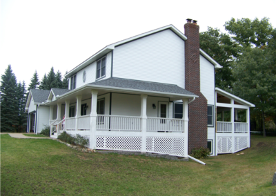 A white two-story house with a brick chimney, gray roofing, and large wraparound porch sits on a grassy hill, surrounded by trees and well-maintained gutters on a cloudy day.
