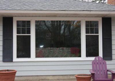 A large, three-panel window with black shutters on a light gray house in Woodbury. Two empty flower pots sit below the window near the gutters, and a purple Adirondack chair is on the right. Trees and furniture are reflected in the glass.