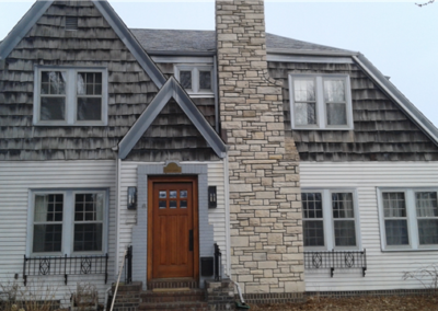 Two-story house with weathered wood shingles above, white siding below, a tall stone chimney, a wooden front door with windows, and several multi-pane windows that add classic charm.