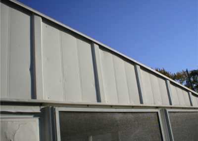 A close-up view of a white metal building exterior with vertical panel siding and quality roofing under a clear blue sky; part of a screened area is visible at the bottom.