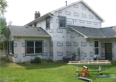 A house under renovation with new siding installation, insulation wrap on the exterior walls, construction materials, and a red sawhorse in the grassy yard. Some windows and landscaping are also visible.