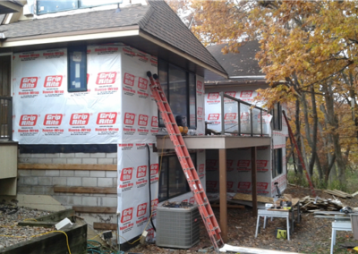 A house under construction with Grip-Rite house wrap on the exterior walls, a red ladder for roofing work, scattered tools and materials on the ground and porch, surrounded by autumn trees.