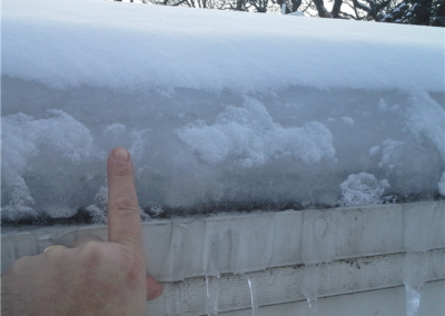 A hand with an extended finger points to a thick layer of snow and ice on the edge of a rooftop, with icicles hanging from the gutters and trees visible in the background.