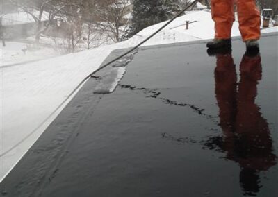 A person in an orange suit uses a pressure washer to clean ice or snow from a roof and gutters, water and reflections glistening on the surface; snowy trees and houses in Woodbury are visible through frosty windows in the background.