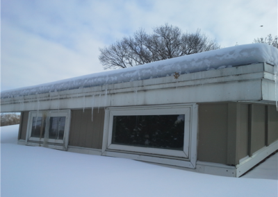 A building in Woodbury is partially buried in snow, with long icicles hanging from the gutters and windows. Bare trees and a cloudy sky are visible in the background.