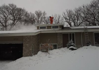 A person in an orange suit is clearing snow from the roof of a house with a snow blower on a snowy day. A ladder leans against the roofing, and bare trees are visible in the background.