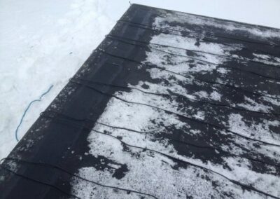 Black heating cables are laid out in a zigzag pattern on a snow-covered roof, helping to melt snow and prevent ice buildup—a smart Roofing solution. Snow is piled up alongside the roof.