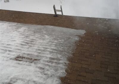 View from a snowy rooftop in Woodbury shows a section partially cleared of snow. A ladder leans against the roof edge near the windows, and a car is parked on a snow-covered street in the background.