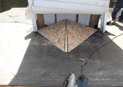 A rooftop under repair in Woodbury features a dormer with exposed plywood forming a triangular pattern at its base, framed by black roofing felt, windows awaiting installation, and construction tools scattered in the foreground.