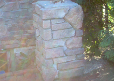 A stone pillar made of irregular, stacked stones stands outdoors next to a stone wall and windows, with some greenery in the background. Sunlight creates a soft glow and lens flare across the image, evoking a peaceful Woodbury setting.