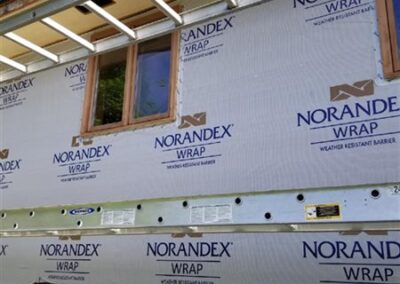 A house under construction with Norandex wrap covering the exterior walls, exposed framing above the windows, and some windows already installed—an example of professional general contracting. A person’s hand is visible at the bottom left corner.