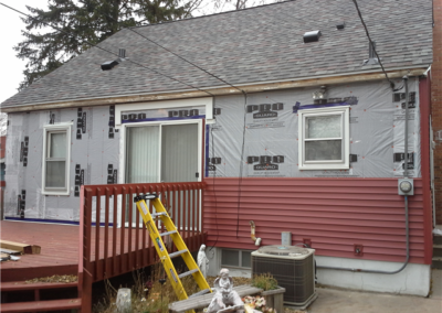 A house exterior in Woodbury under renovation, with new red siding partially installed. The upper wall is covered in protective wrap, a yellow ladder leans on the deck, and roofing tools and general contracting materials are scattered nearby.