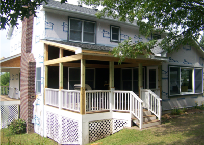 A two-story house with windows, a partially constructed wooden porch, white railing, and lattice skirting. The exterior walls are covered with house wrap, and construction materials are visible on the porch.